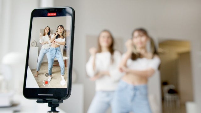 Registrazione video di due ragazze in uno studio di produzione, con uno smartphone che riprende la scena, evidenziando contenuti creativi e di moda.