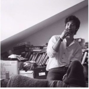 Un uomo seduto in un ambiente di lavoro con scaffali pieni di libri e materiali, in un'atmosfera creativa e informale, riflettendo il concept di The Usual NeXt.