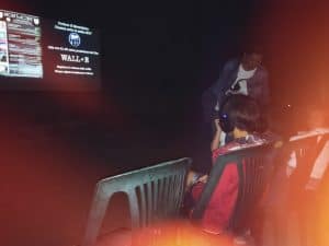 Proiezione di un film al cinema all'aperto Montalcino, pubblico seduto con cuffie, atmosfera notturna, evento culturale in Italia, promozione culturale, giovane spettatore con cuffie.