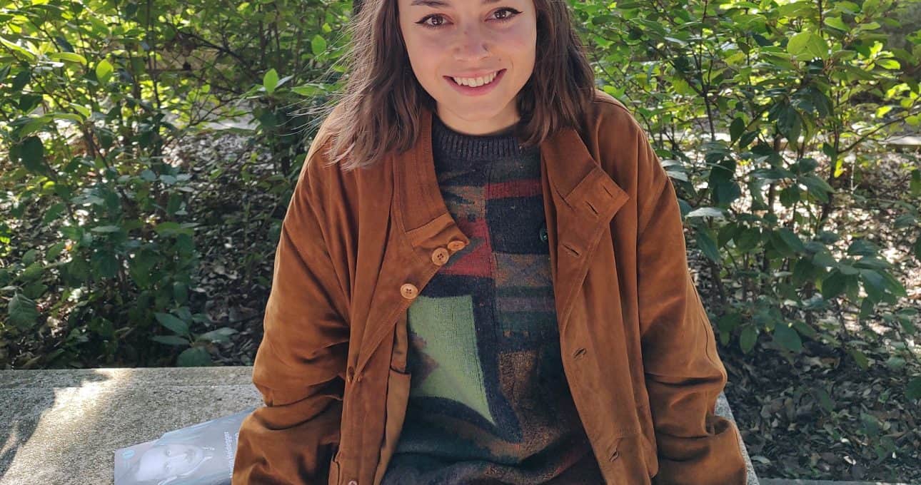 Femminile giovane seduta su panca in parco con giacca marrone e maglione colorato, natura e alberi sullo sfondo, ambiente outdoor, stile casual, lifestyle, atmosfera rilassante, urban outdoor, ritratto donna.