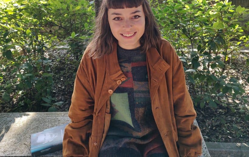 Femminile giovane seduta su panca in parco con giacca marrone e maglione colorato, natura e alberi sullo sfondo, ambiente outdoor, stile casual, lifestyle, atmosfera rilassante, urban outdoor, ritratto donna.