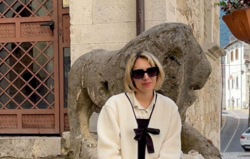 Marta seduta davanti a Stone Lion statue in un vicolo di un borgo storico italiano con edifici antichi e montagne sullo sfondo, abbigliamento elegante e occhiali da sole, atmosfera di charme e tradizione locale.