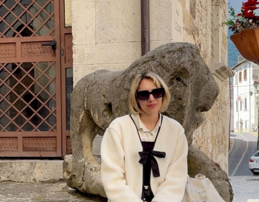 Marta seduta davanti a Stone Lion statue in un vicolo di un borgo storico italiano con edifici antichi e montagne sullo sfondo, abbigliamento elegante e occhiali da sole, atmosfera di charme e tradizione locale.